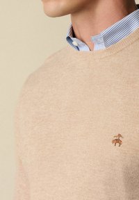 Maglione lavorato a maglia beige con scollo a girocollo, caratterizzato da un logo ricamato di dimensioni ridotte. Indossato sopra una camicia a colletto blu chiaro e bianca a righe.