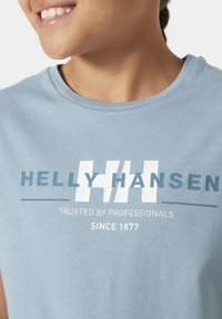 Camiseta de algodón azul claro con cuello redondo, gran logo de "Helly Hansen" en azul oscuro y blanco, con un acabado texturizado y texto impreso debajo.