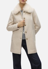 Manteau en laine beige avec un col en fausse fourrure, doté de boutons dorés, de poches latérales et d'une coupe ajustée. Porté sur un pull gris et un jean foncé.