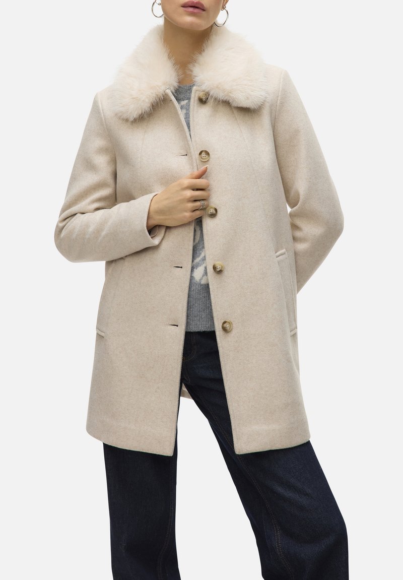 Manteau en laine beige avec un col en fausse fourrure, doté de boutons dorés, de poches latérales et d'une coupe ajustée. Porté sur un pull gris et un jean foncé.
