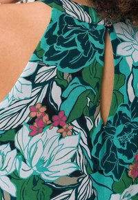 Robe fleurie en tissu vert et bleu sarcelle avec des motifs de fleurs blanches et roses. Dos avec ouverture en forme de goutte et finition douce et texturée.