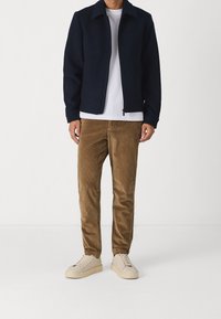 Giacca navy con zip e collo a risvolto, abbinata a pantaloni in velluto a coste color sabbia e sneaker chiare, caratterizzata da una texture liscia e linee pulite.