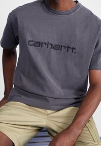 Grijze katoenen T-shirt met ronde hals en geborduurd "carhartt." logo in donkergrijs, gecombineerd met beige shorts. Gladde textuur, casual pasvorm.