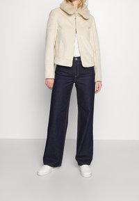 Giacca in finta pelliccia color crema con colletto, chiusura a zip e tasche laterali, abbinata a jeans in denim scuro a vita alta e sneakers bianche.