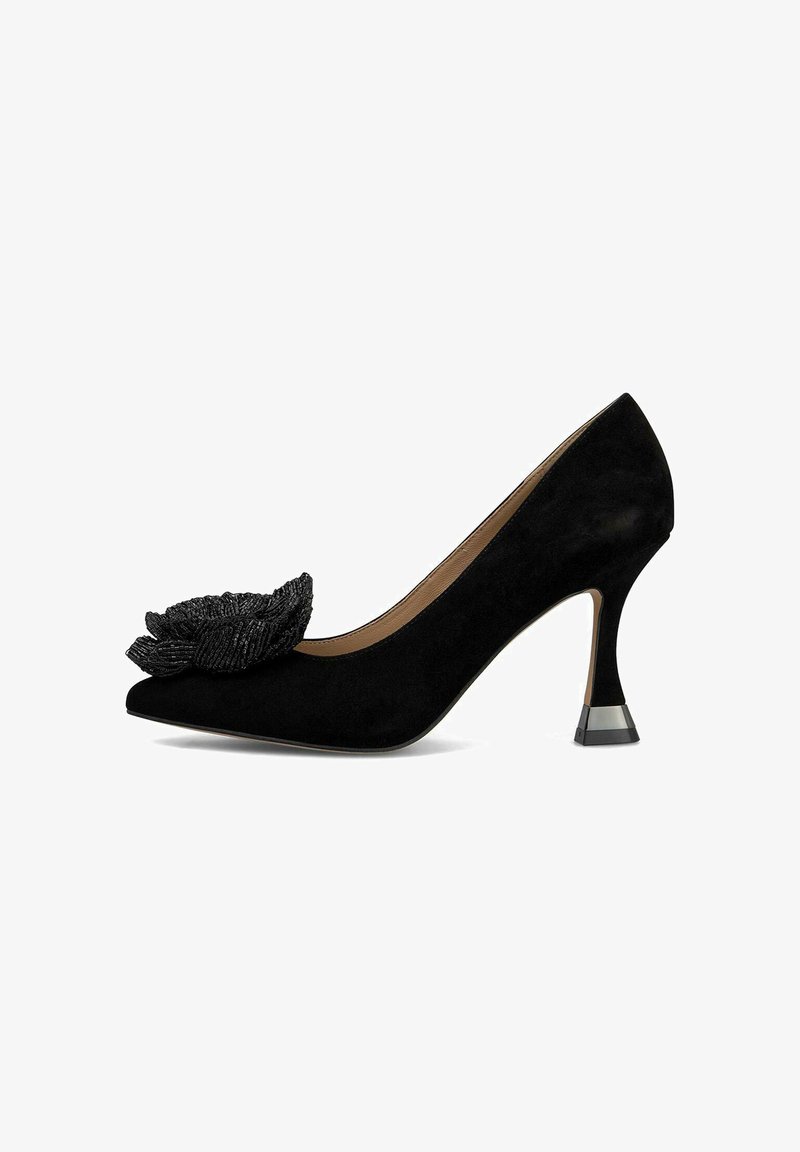 Zapato de tacón alto de ante negro con punta afilada, que presenta un acento de flor texturizada negra en la parte frontal y un tacón transparente en bloque.