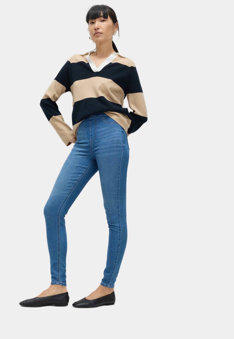 Kvinde stående med hænderne på hofterne, iført beige og sort stribet sweater, blå skinny jeans og sorte flade sko, foran en ensfarvet baggrund.