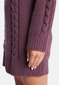 Lähikuva kädestä, jossa on vaaleanpunaiset kynnet, lepäämässä mauve-värisen neulottu mekko, jonka hihoissa on villatikkikuvioita.