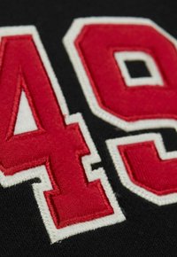 Schwarzer Textil mit großen roten und weißen Applikationszahlen "49" und "0". Die Nähte sind sichtbar, was die Struktur und Details hervorhebt.