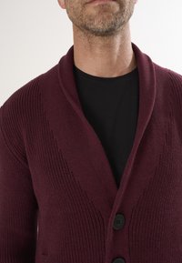 Burgundi värvi kardigan, millel on šalli kaelus, ribakonstruktsioon ja ees kolm musta nööpi. Kantud musta särgi peal.