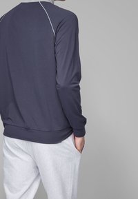 Person wearing a dark long-sleeve shirt with white piping and light gray textured pants, standing with one hand in pocket against gray background.