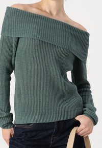 Maglione a spalla scoperta in maglia rib rib verde teal, con scollo ampio e polsini aderenti. Abbinato a jeans scuri e una borsa chiara.