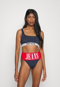 Námornícky bikini top s bielym pásikom s nápisom "TOMMY JEANS," v kombinácii s vysokými červenými spodkami so širokým bielym nápisom "JEANS." Hladká textúra.