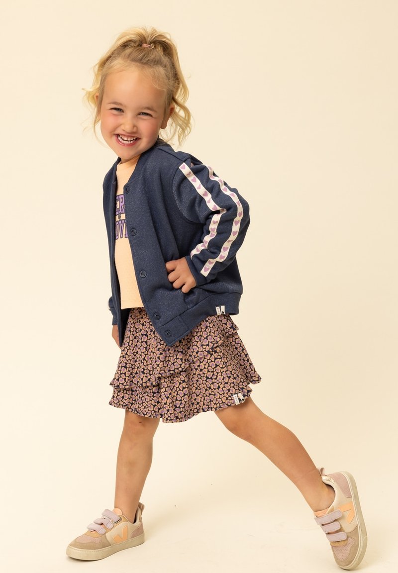 Navyblauwe cardigan met strepen met meerkleurige hartjespatronen, licht perzikkleurig shirt, gelaagde bloemige rokken met ruches en beige sneakers met klittenband.