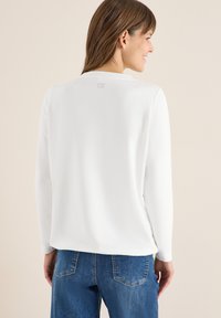 Witte langeblaffen top met een ronde halslijn. Zachte textuur met een subtiele logo op de achterkant. In combinatie met blauwe denim jeans.