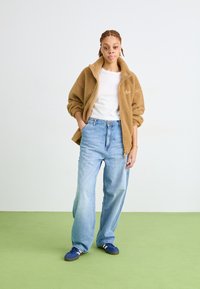 Jachetă din fleece bej, tricou alb, blugi largi de culoare albastru deschis și adidași albastre. Material neted, croială lejeră și fermoar frontal pe jachetă.