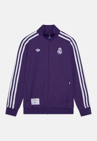 Lila zip-up jacka med vita ränder, med Adidas och Real Madrid-logotyper. Tillverkad av mjukt tyg med ribbad mudd och nederkant.