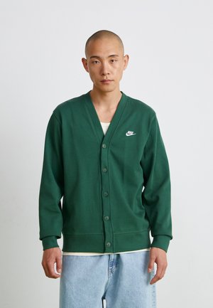 Cardigan vert en tissu doux, avec un col en V, fermeture à boutons et un logo Nike blanc sur la poitrine gauche. Coupe décontractée.