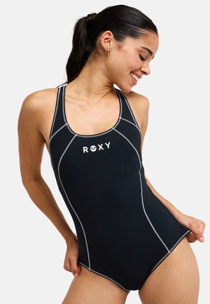 Femme souriante aux cheveux foncés attachés en queue de cheval, portant un maillot de bain une-pièce noir Roxy avec des coutures blanches sur fond blanc.