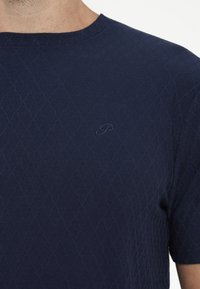 Chemise bleu foncé à manches courtes avec un motif subtil en forme de losange et une petite lettre P brodée sur la poitrine, portée par une personne.