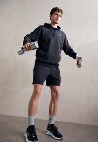 Ung mand i sort hættetrøje og shorts, der løfter sølvfarvede håndvægte i et neutralt indendørs rum, iført sorte sneakers og grå strømper.