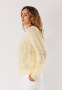 Donna con capelli ondulati che indossa un maglione a maglia giallo chiaro e pantaloni bianchi, in piedi di lato con le mani nelle tasche su uno sfondo neutro.