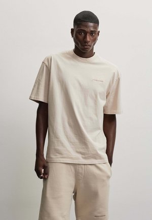 Junger Mann trägt ein hellbeiges oversized T-Shirt und passende Shorts, steht vor einem schlichten hellen Hintergrund.