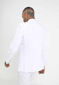 Chaqueta de traje blanca con un acabado de tejido suave, una abertura en la parte trasera y detalles sutiles en los puños. Modelo mostrado desde atrás.