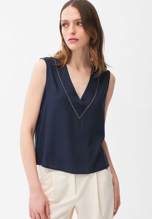 Jonge vrouw met schouderlang bruin haar, gekleed in een mouwloos marineblauw V-hals topje en crèmekleurige wijde broekspijpen, kijkt naar links.