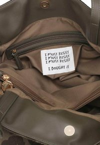 Interno di una borsa verde oliva con un'etichetta di stoffa su cui è scritto: "DEVO RESISTERE" ripetuto, seguito da "L'HO COMPRATA". Include una tasca con cerniera.