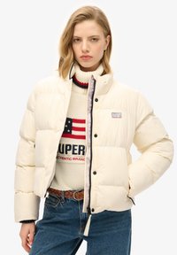 Hvid pufferjakke med en cropped design, der har sorte knapper og et logo patch. Båret over en cremefarvet sweater med røde og blå accenter.