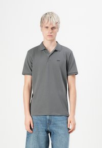 Jeune personne avec les cheveux blonds courts, portant un polo gris de GAP et un jean bleu, se tenant contre un fond blanc uni.