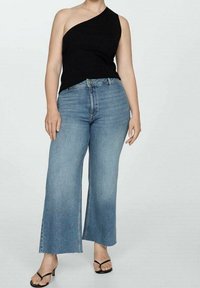 Top negro de un solo hombro con textura acanalada combinado con vaqueros de pierna ancha en azul claro. Los vaqueros tienen un dobladillo sin rematar, y la modelo lleva sandalias negras de tiras.