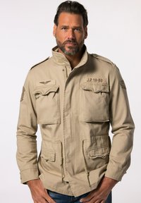 Giacca militare beige con collo alto, due tasche sul petto e cordini regolabili in vita. Tessuto testurizzato con spalline.