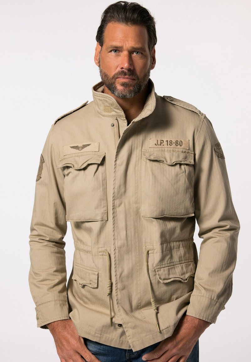 Giacca militare beige con collo alto, due tasche sul petto e cordini regolabili in vita. Tessuto testurizzato con spalline.