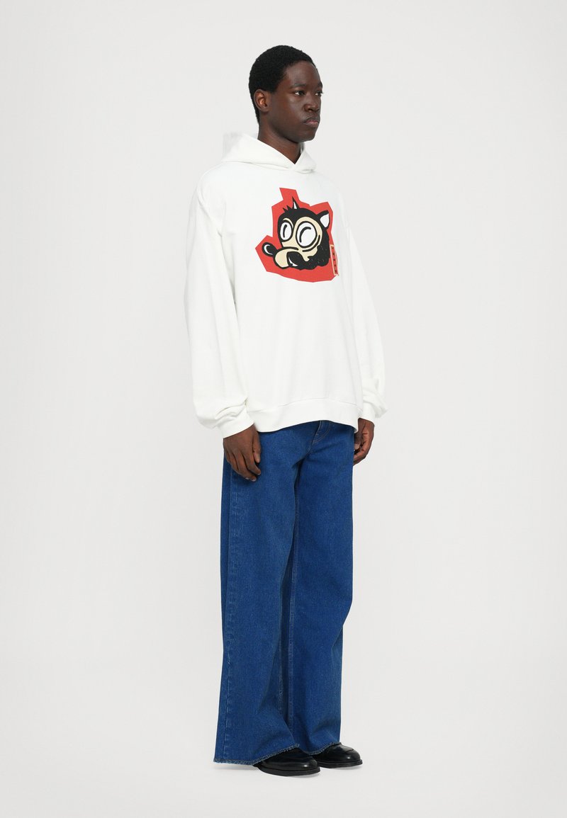 Hvid hættetrøje med en farverig grafik, der viser en tegneseriefigur, parret med løstsiddende blå denim jeans og sorte sko.