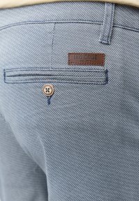 Gros plan sur la poche arrière d’un pantalon texturé bleu avec bouton et étiquette en cuir marron portant l’inscription "INDICODE EST. 1995."