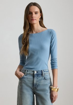 Frau mit langen braunen Haaren, die ein hellblaues, geripptes Oberteil, helle Jeans, ein massives goldenes Armband und blaue Ohrringe vor einem einfarbigen Hintergrund trägt.