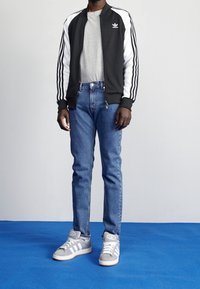 Černobílá bunda na zip, šedé tričko, modré džíny a šedé tenisky s bílými pruhy, stojící na modrém povrchu.