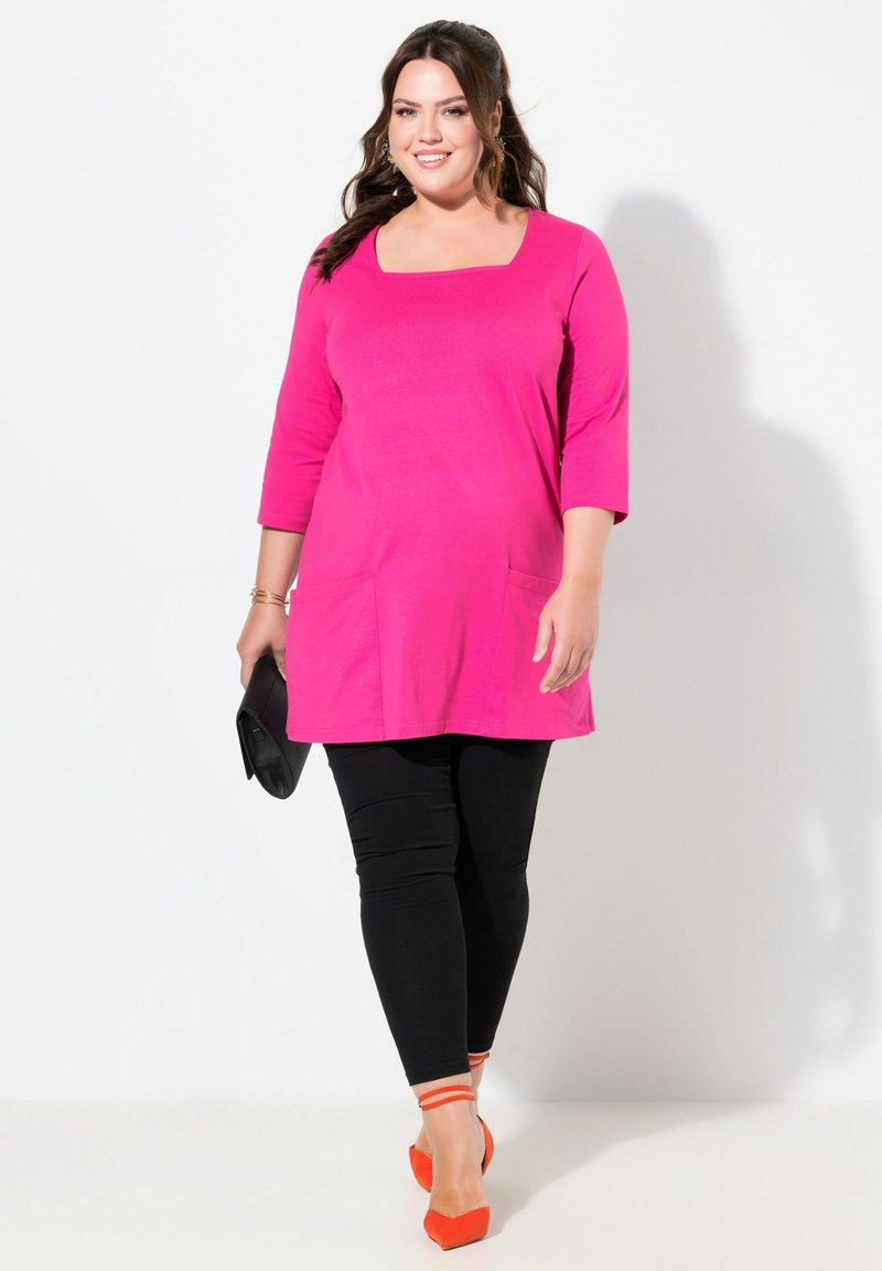 Rosa Tunika-Top mit quadratischem Ausschnitt und zwei Vordertaschen, kombiniert mit schwarzen Leggings und orangefarbenen Absatzschuhen mit Knöchelriemen.