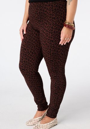Leggings met een bruin-zwart luipaardprint, gedragen met witte gevlochten platte schoenen, gouden armbanden, ringen en een gedeeltelijke tatoeage zichtbaar op de linkerarm.