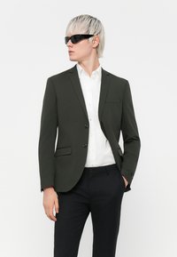 Mørkegrøn blazer med to knapper, en enkelt brystlomme, båret over en hvid skjorte, parret med sorte bukser, og accessoriseret med sorte solbriller.