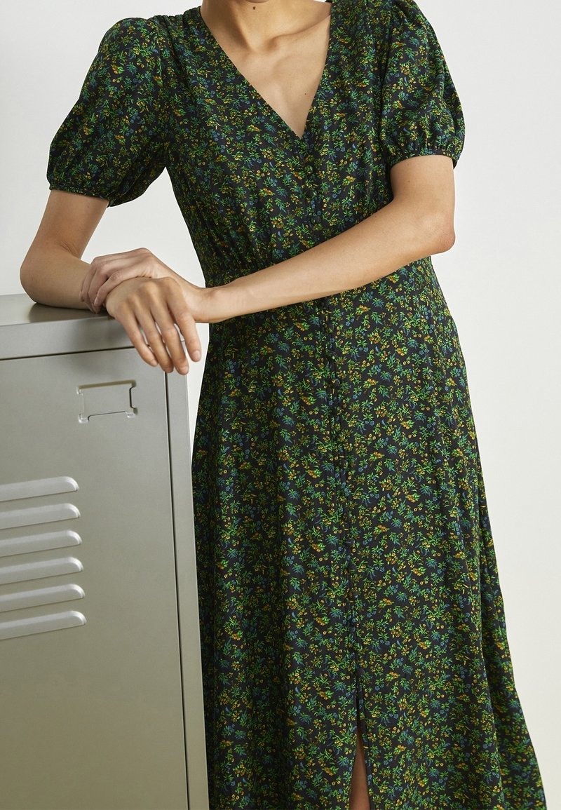 Personne en robe fleurie vert foncé à manches courtes bouffantes, appuyée sur un casier en métal, seulement le haut du corps et le torse visibles.