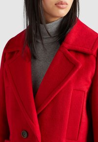 Femme portant un manteau en laine rouge vif par-dessus un pull à col roulé gris côtelé, mettant en valeur le grand revers et les détails de poche du manteau.