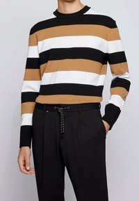 Pull rayé noir, blanc et marron clair avec un col rond, en maille texturée, associé à un pantalon noir avec une taille à cordon.