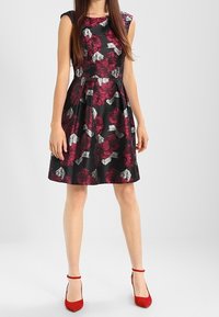 Černé květinové šaty s vínovými a stříbrnými květinovými vzory, upnutý korzet, rozšířená sukně a rukávy do půlky. Doplněné červenými lodičkami.