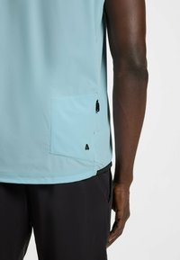 Light blue sleeveless top with a pocket, featuring a zipper and branding detail. Fabric appears smooth with a matte finish.