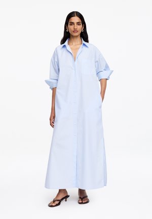 Femme portant une longue robe-chemise bleu clair boutonnée, manches retroussées et sandales à talons noires à bouts ouverts sur fond blanc.