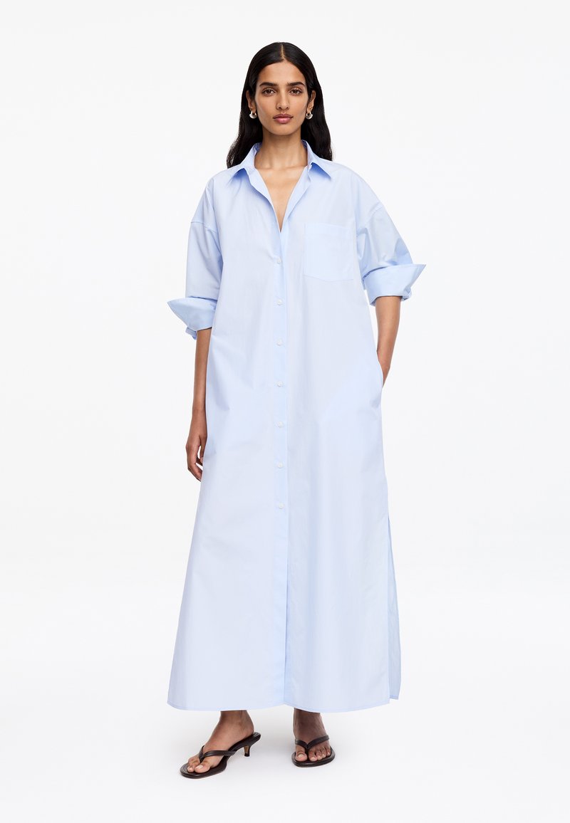 Femme portant une longue robe-chemise bleu clair boutonnée, manches retroussées et sandales à talons noires à bouts ouverts sur fond blanc.