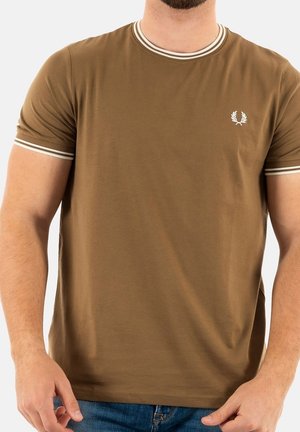 Mężczyzna w brązowej koszulce z krótkim rękawem, z białym paskowanym kołnierzykiem i mankietami, z małym białym logo wawrzynu na lewej piersi, oraz w niebieskich dżinsach.