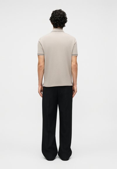 Άνδρας με σγουρά μαλλιά, γυρισμένος προς τα πίσω, φοράει μπεζ κοντομάνικο πόλο και χαλαρά μαύρα παντελόνια, μπροστά σε απλό φόντο.